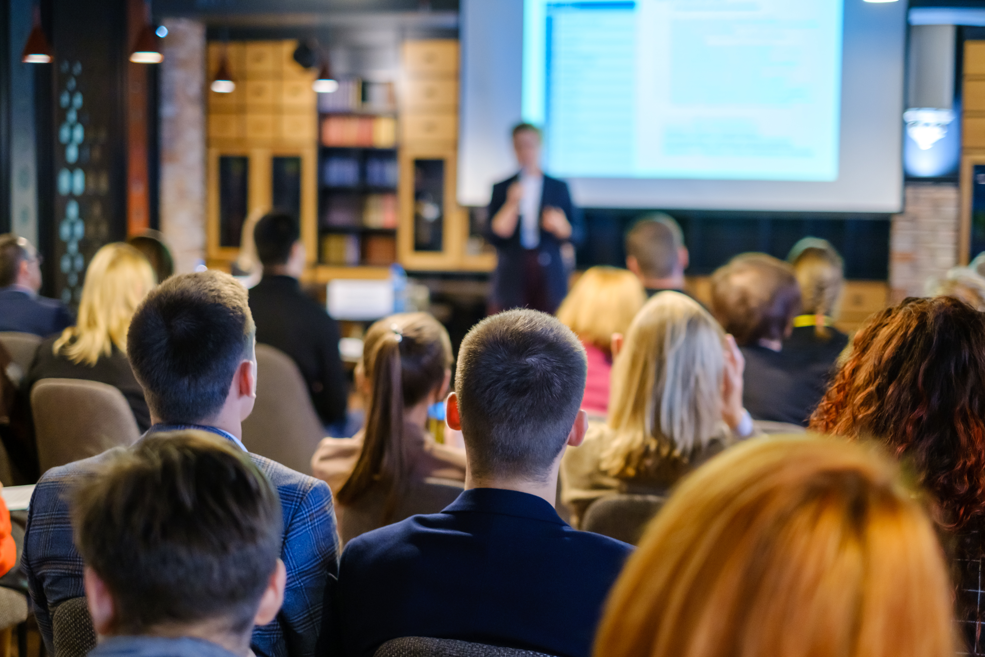 Audience attending a business presentation or conference, watching a speaker in front of a large projection screen.