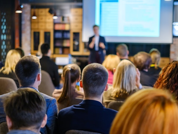 Audience attending a business presentation or conference, watching a speaker in front of a large projection screen.