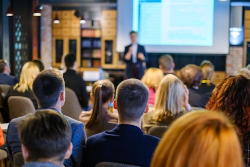 Audience attending a business presentation or conference, watching a speaker in front of a large projection screen.