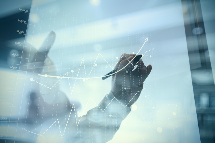 Hand drawing an upward-trending line on a transparent screen overlaid with financial graphs, symbolising growth and success.