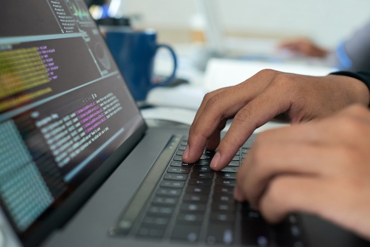 Close-up of hands typing on a laptop keyboard with lines of colourful code displayed on the screen.