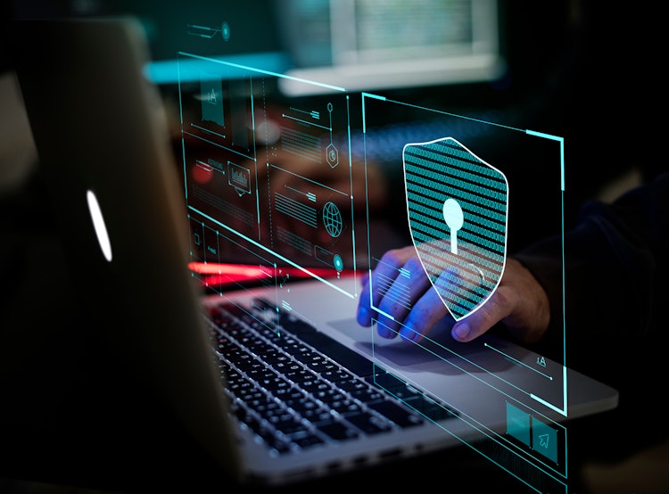Person working on a laptop with holographic cybersecurity interface overlays, including a digital shield and lock icon representing data protection.