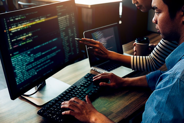 Two programmers collaborating at a desk, reviewing code on dual monitors and a laptop while discussing debugging.