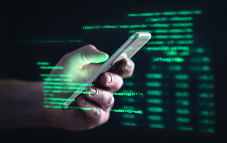Close-up of a hand holding a smartphone with green glowing code and digital data projected in the background, symbolizing cybersecurity or mobile hacking.