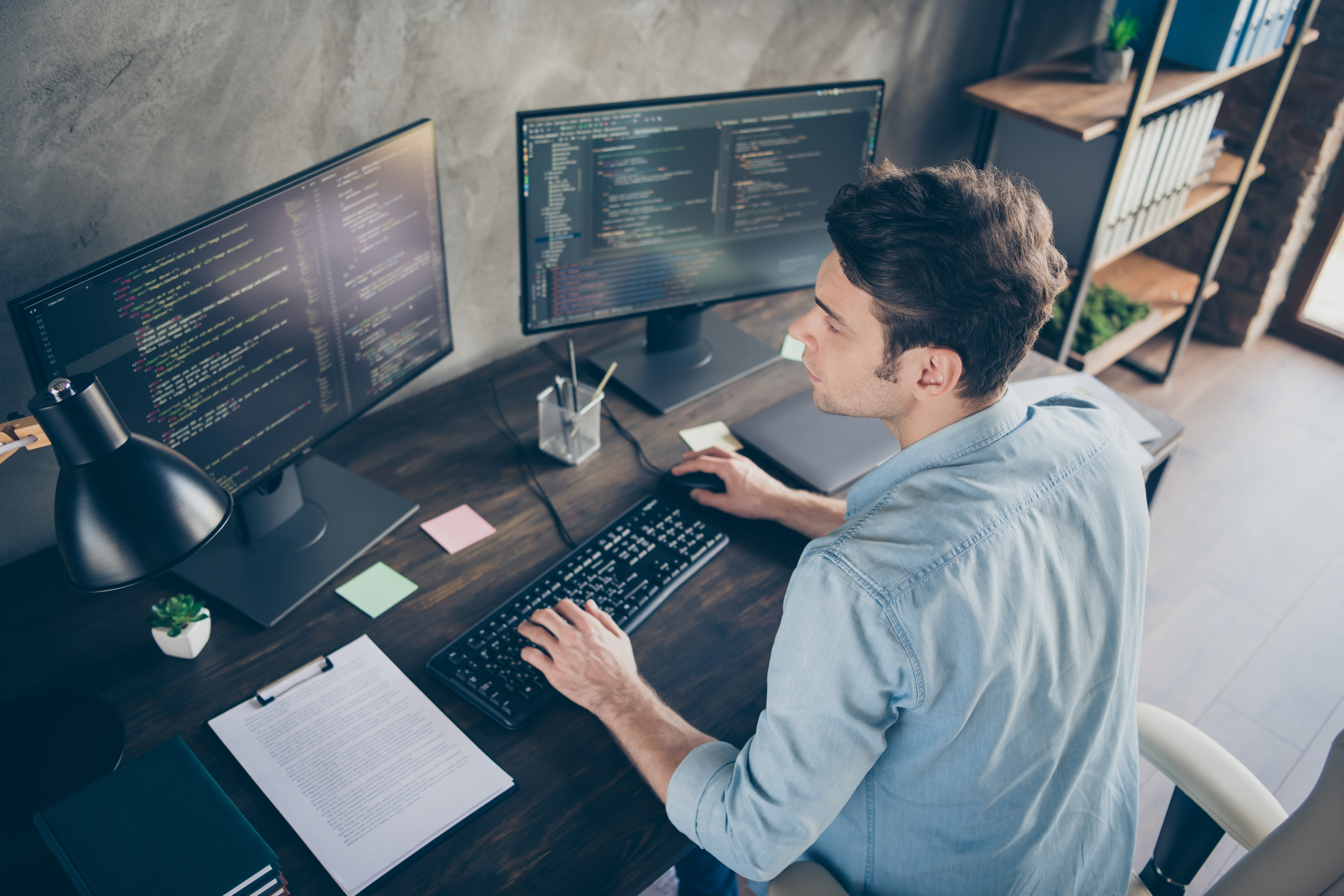 Software developer working at a desk with dual monitors displaying code in a modern home office setup.