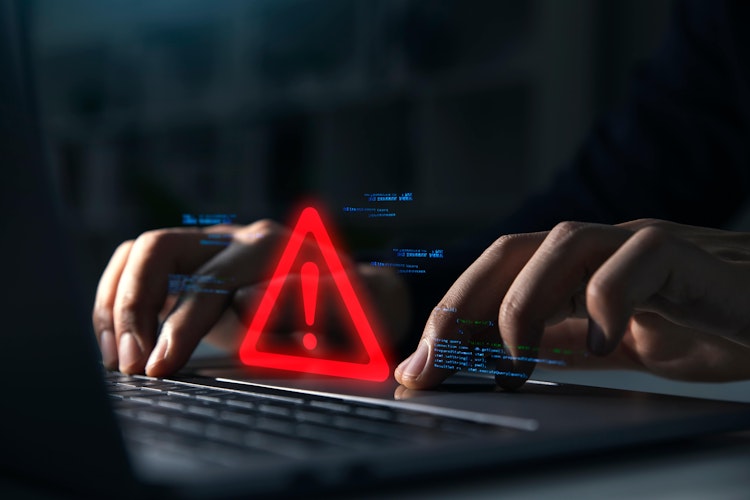 Close-up of hands typing on a laptop with a glowing red warning triangle and exclamation mark, symbolising a cybersecurity threat or system error.