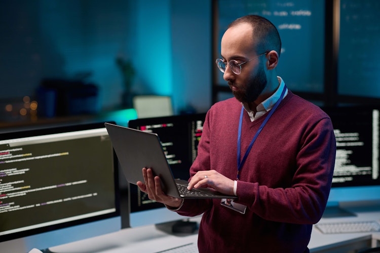 IT professional in a modern office working on a laptop with multiple monitors displaying code in the background.