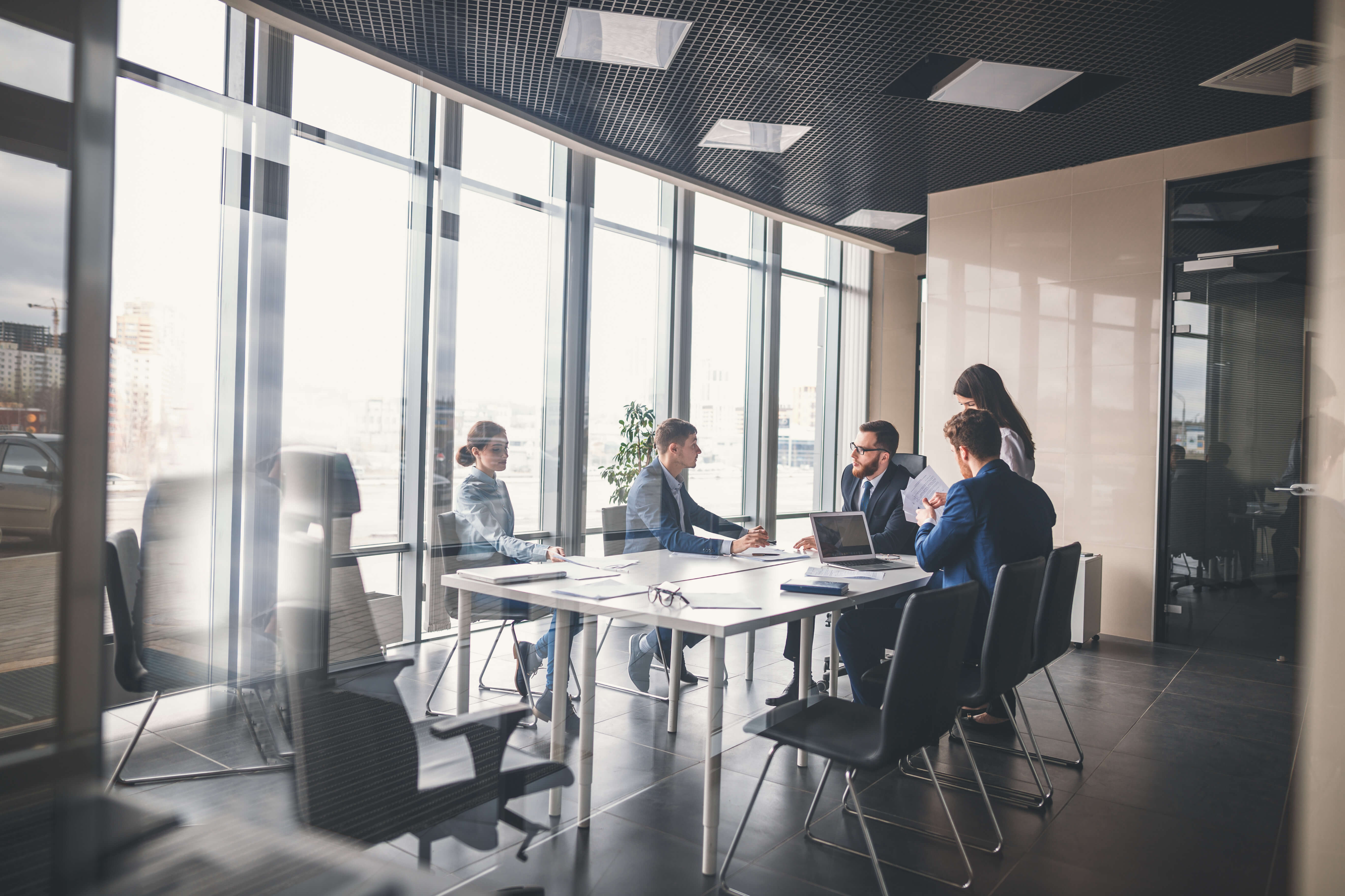 Business team in formal attire having a meeting in a modern glass-walled conference room with laptops and documents on the table.