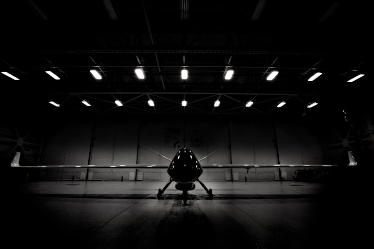Dramatic black and white photo of a drone or aircraft silhouetted inside a dimly lit hangar under overhead lights.