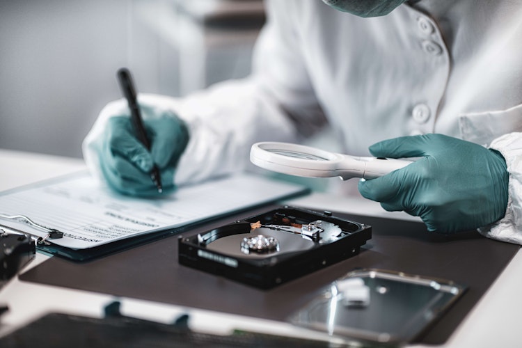 Forensic investigator examining a disassembled hard drive with a magnifying glass while taking notes.