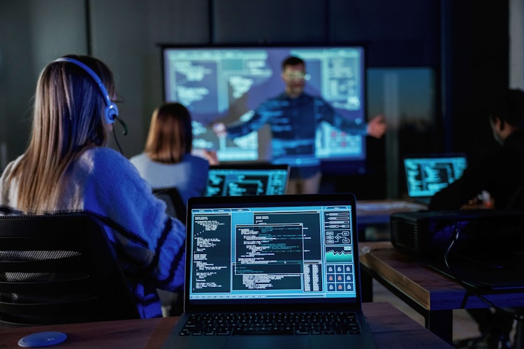 Cybersecurity training session with participants using laptops displaying code while an instructor presents on a large screen.