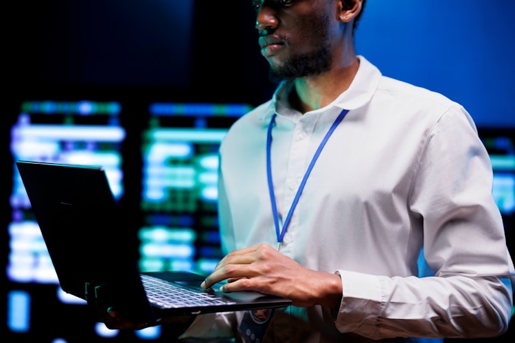 Cybersecurity professional working on a laptop in a control room with digital screens in the background.