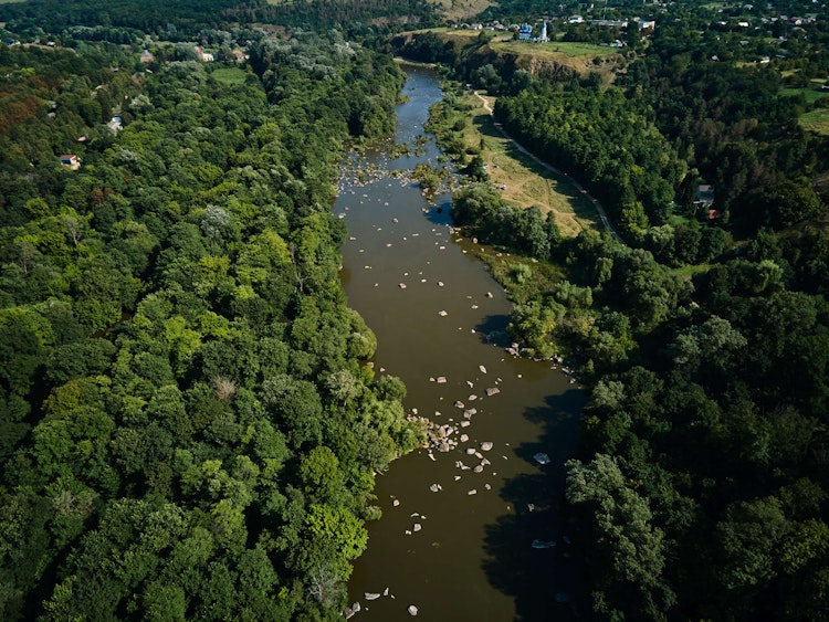 Aerial view of a river winding through dense green forest with scattered rocks and small settlements in the distance.