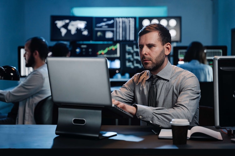 Focused professional working on a laptop in a control room with multiple data and world map screens in the background.