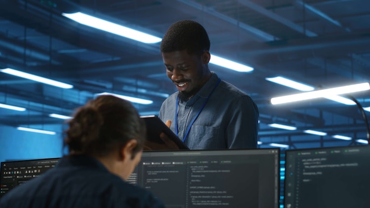 Smiling IT professional reviewing a tablet while colleagues work on coding at computer monitors in a modern office.