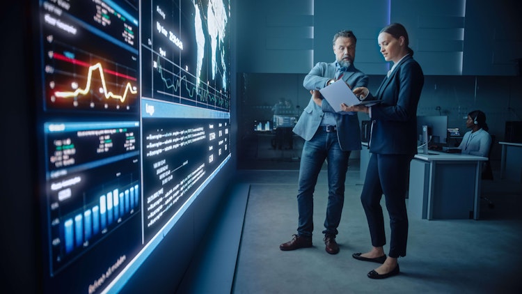 Two business professionals reviewing documents in front of a large digital wall displaying global data, charts, and analytics.