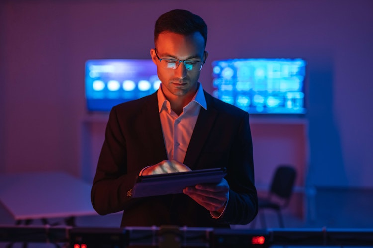 Business professional in a dark room using a tablet, illuminated by blue and red light with digital screens in the background.