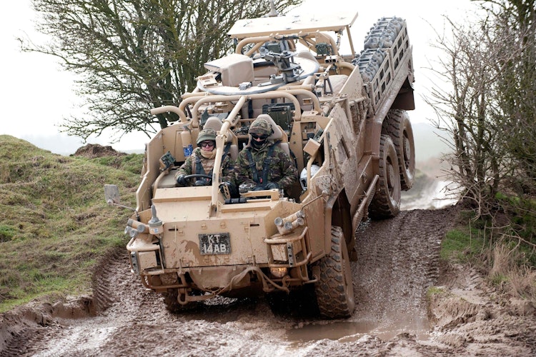 Military personnel driving a camouflaged off-road armoured vehicle through a muddy terrain.