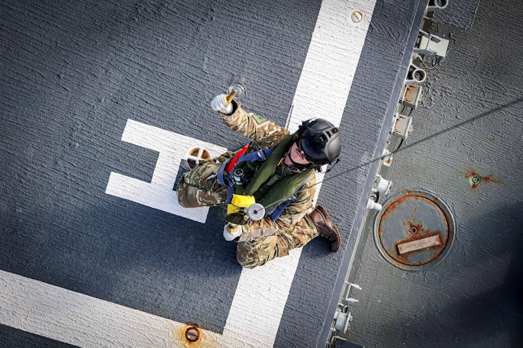 Soldier in camouflage uniform and helmet kneeling on a ship’s deck, holding signalling equipment for aircraft or helicopter operations.