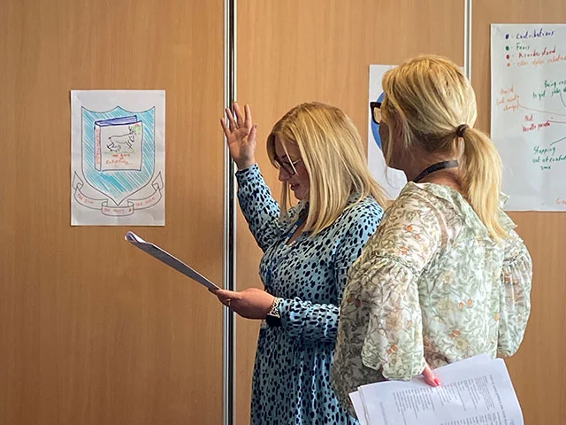 Two women holding papers engaged in a discussion in front of posters on a wooden wall.