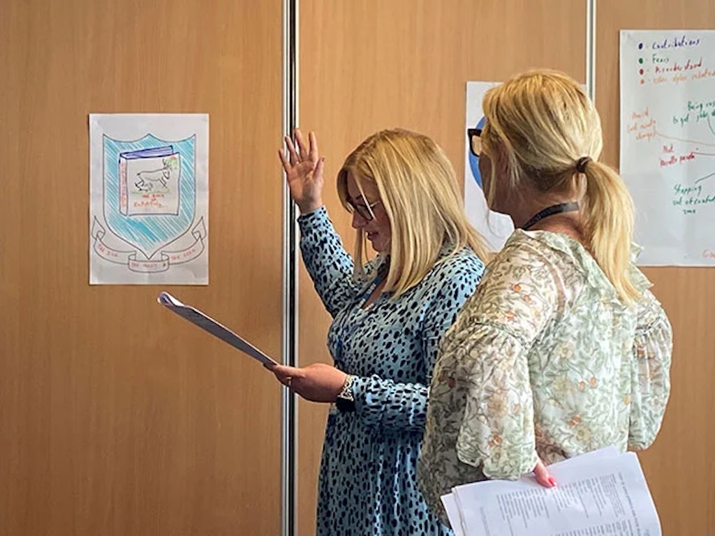 Two women holding papers engaged in a discussion in front of posters on a wooden wall.