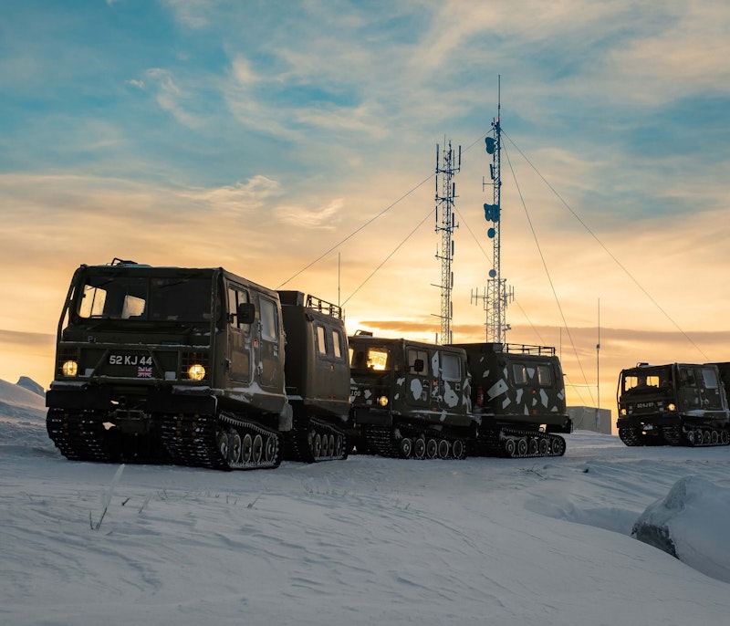 Military tracked vehicles in snowy terrain with communication towers at sunset.