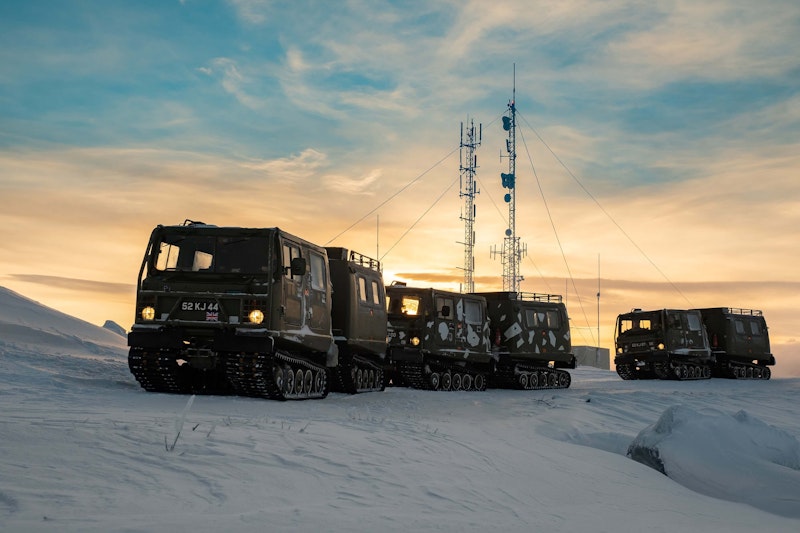 Military tracked vehicles in snowy terrain with communication towers at sunset.