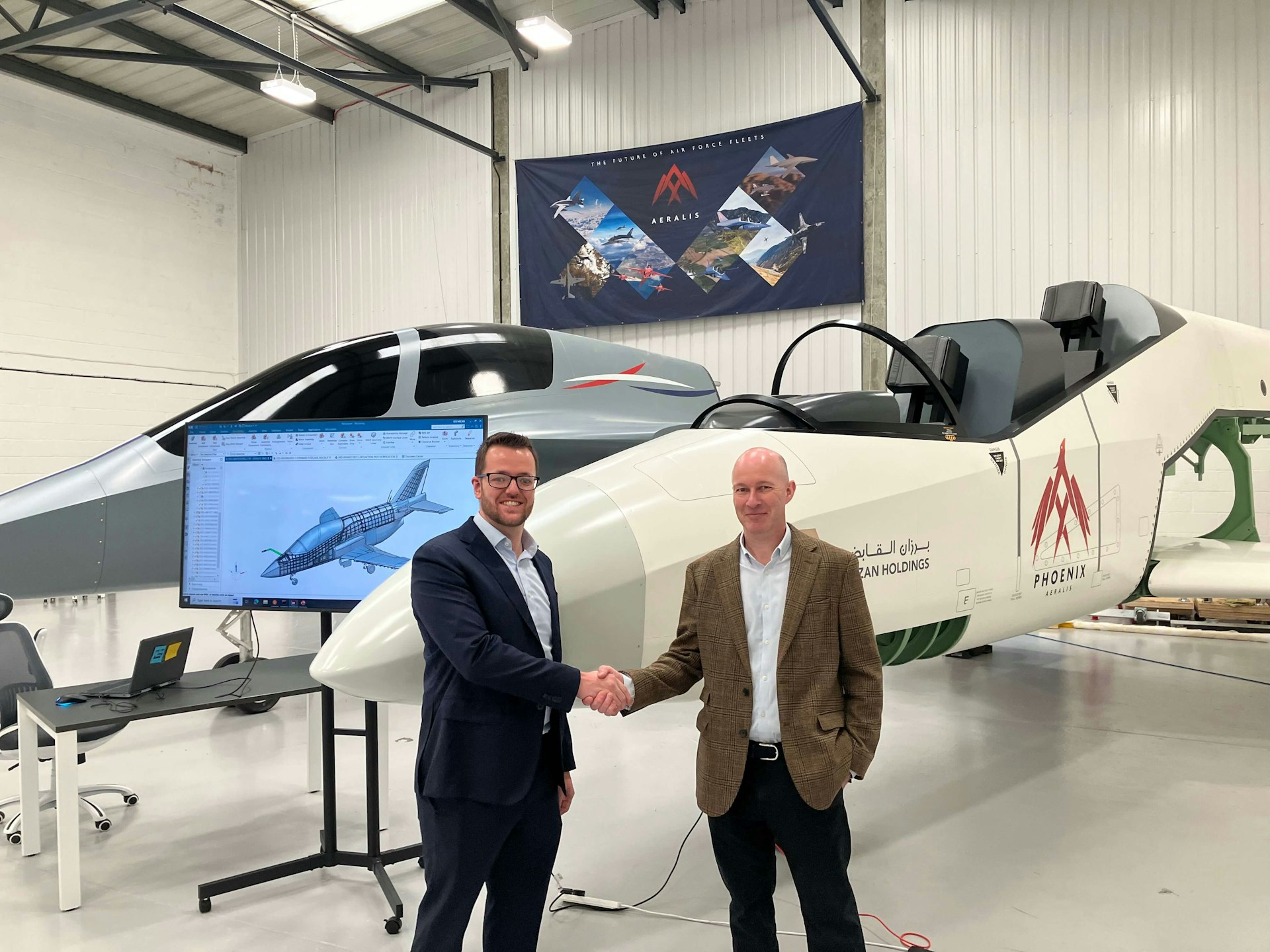 Two men shaking hands in front of a prototype aircraft inside a hangar.