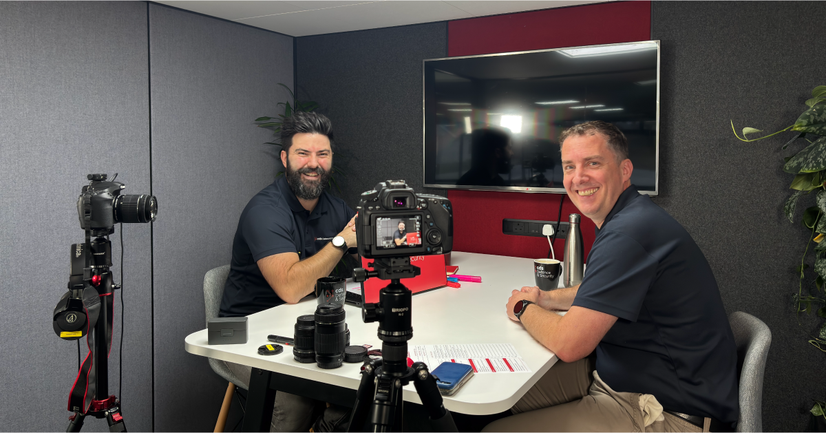 Podcast host Karl Rees and guset Andy Davis smiling infront of 2 DSLR cameras presenting podcast. Tv in the background with two green bushes