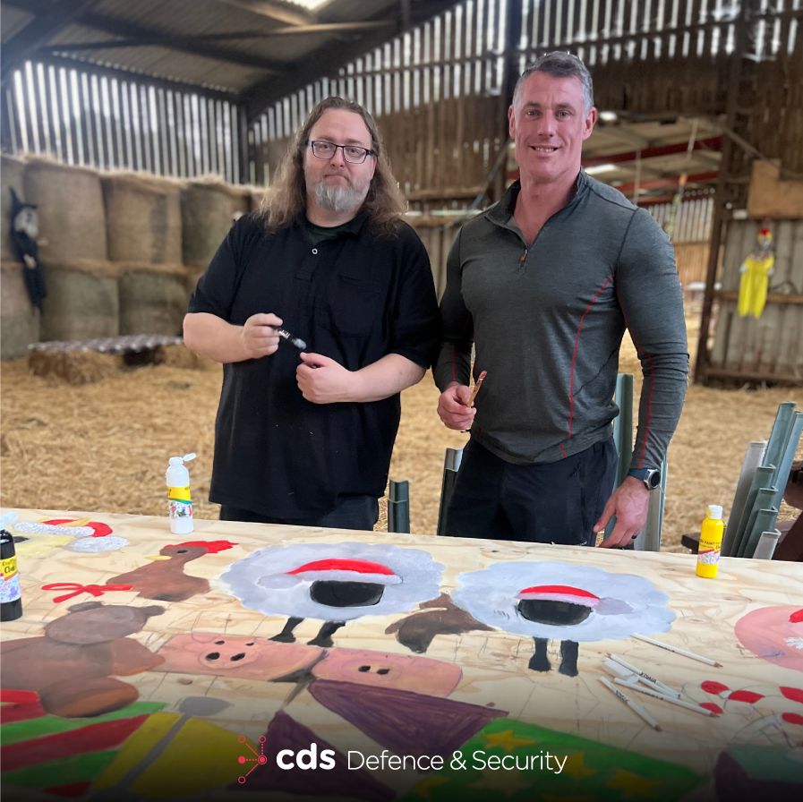 CDS DS employees Austen Shand and Richard Richardson stand in a barn, holding paint brushes, infront of a christmas mural