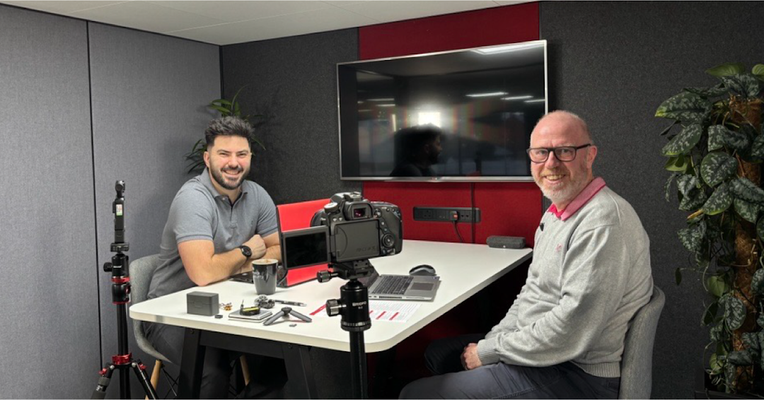 Two CDS DS employees Karl Rees and Graham Hill smiling sat down in a meeting room infront of cameras, ready t orecord a podcast