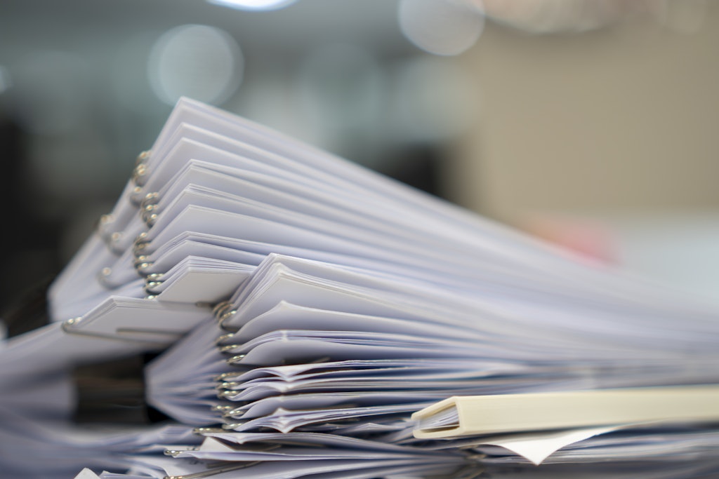 A low angle view focuses on a pile of white documents with metal binding lying on a surface. The office environment suggests a workplace setting, conveying the concept of paperwork, bureaucracy, or administrative tasks. The image could be used in projects about office work, business, and administration. The bright lighting creates a sense of order and precision