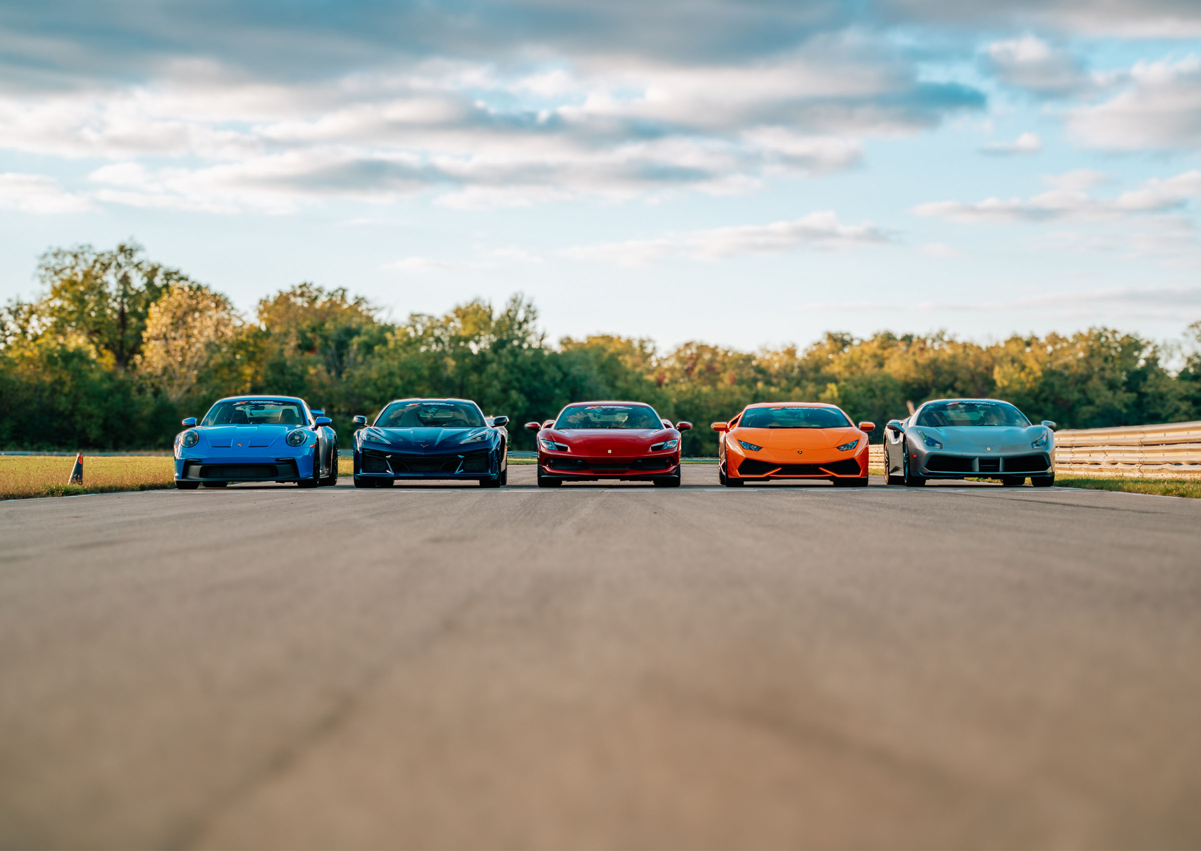 Blue Porsche 911 GT3, Black Corvette C8 Z06, red Ferrari 296 GTB, orange Lamborghini Huracan LP610-4, and Gray Ferrari 488 GTB on the track parked for a photo at Autobahn Country Club.