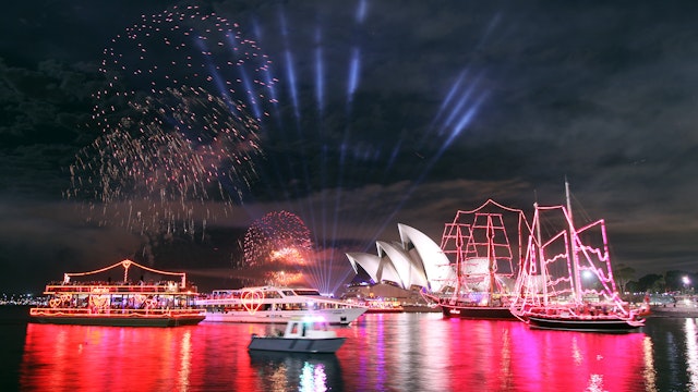 Search lights point into the night sky with illuminated sailing ships and boats on the harbour in front of the Sydney Opera House.