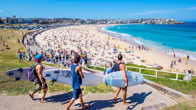 3 surfers walking down the concrete path to the promenade along Bondi Beach, busy with sunbathers on the sand and swimmers in the surf.
