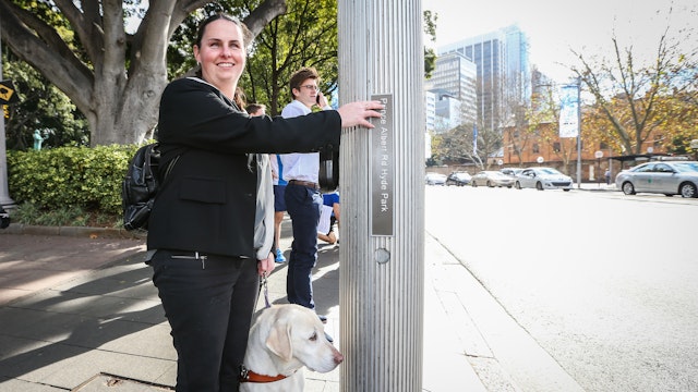 A woman with a guide dog uses a tactile street sign at a pedestrian crossing.