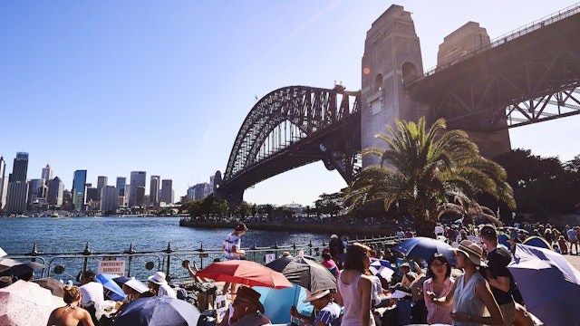 Scores of spectators wait during the day along the northern foreshore of Sydney Harbour with the Sydney Harbour bridge in the background.