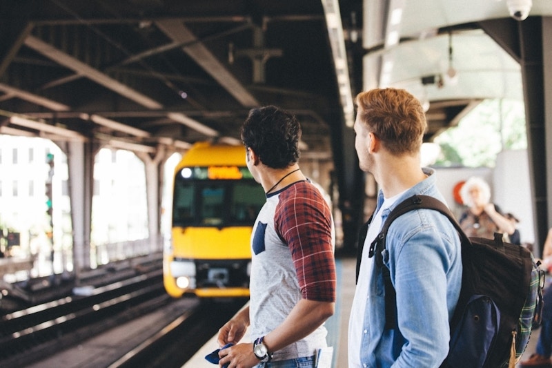 Two people stand on a train platform looking at a yellow train approaching. Other passengers are visible in the background under the station’s canopy.