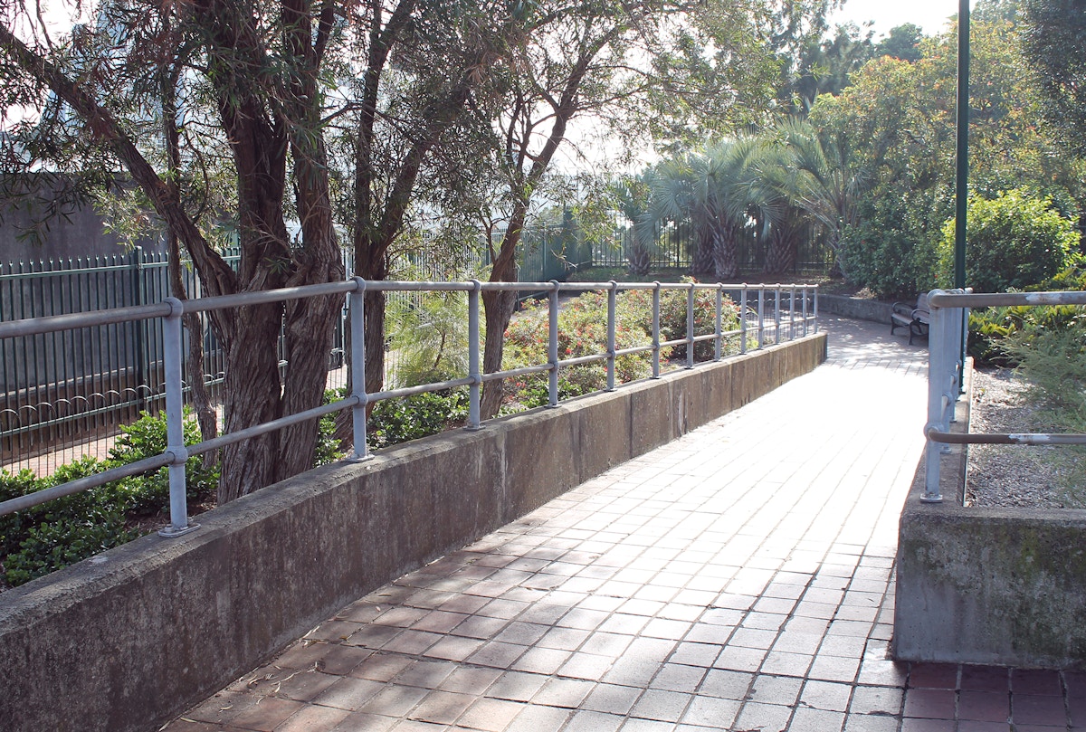 Paved pathway with metal railings, lined by trees and shrubs, curves to the right in a sunlit garden area.