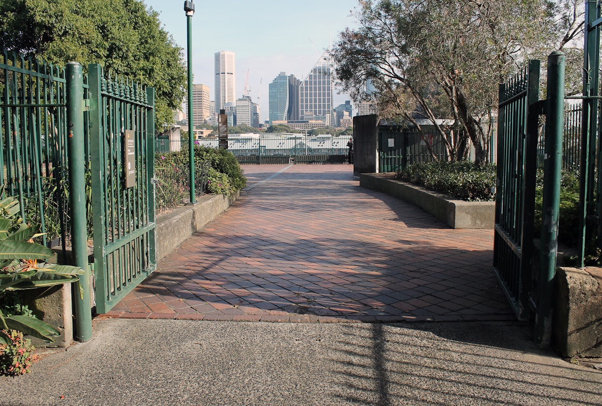 Open green iron gates lead to a brick-paved walkway with city buildings visible in the background and trees on both sides.