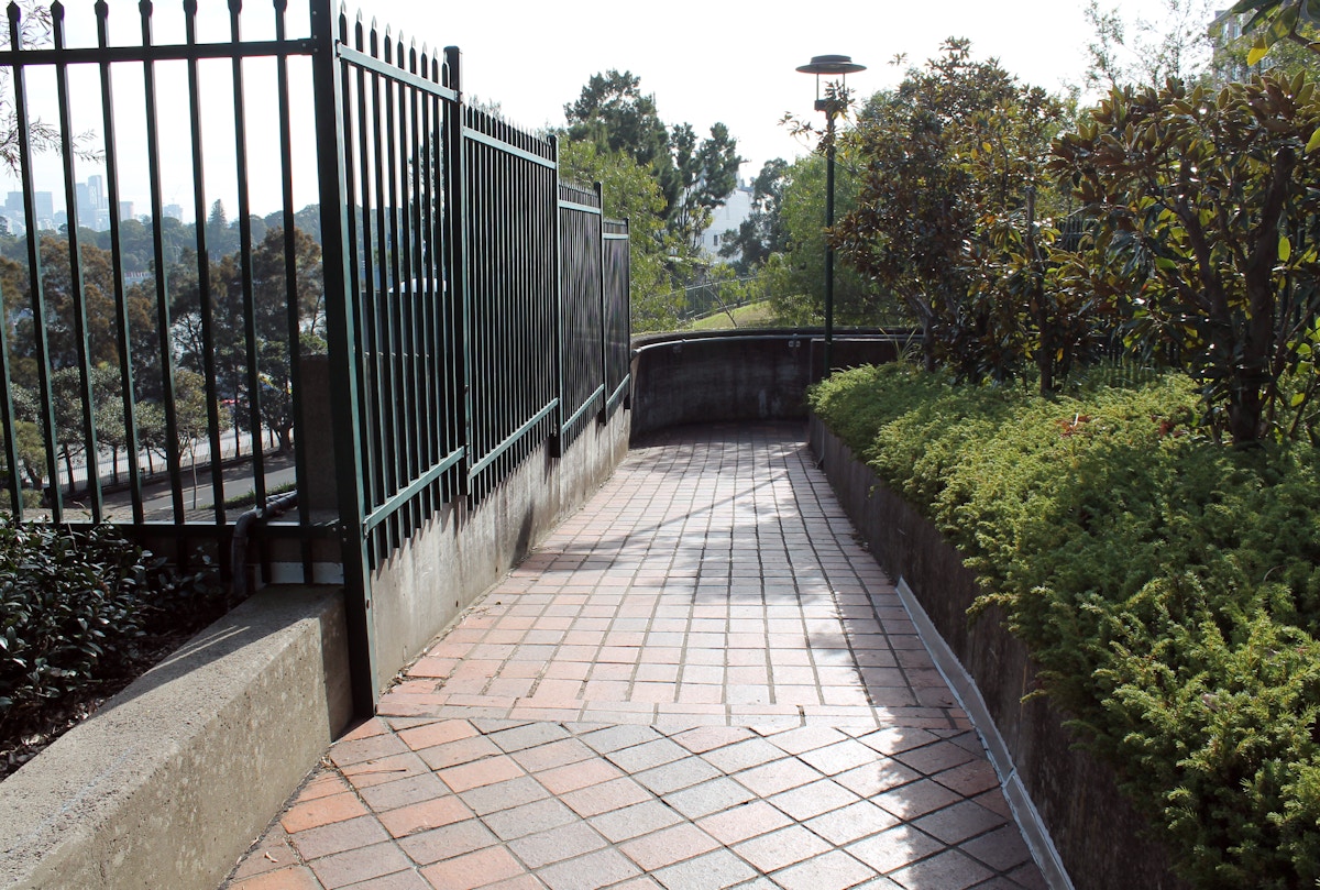 A paved walkway curves alongside a metal fence and green bushes, with trees and a lamp post visible in the background.