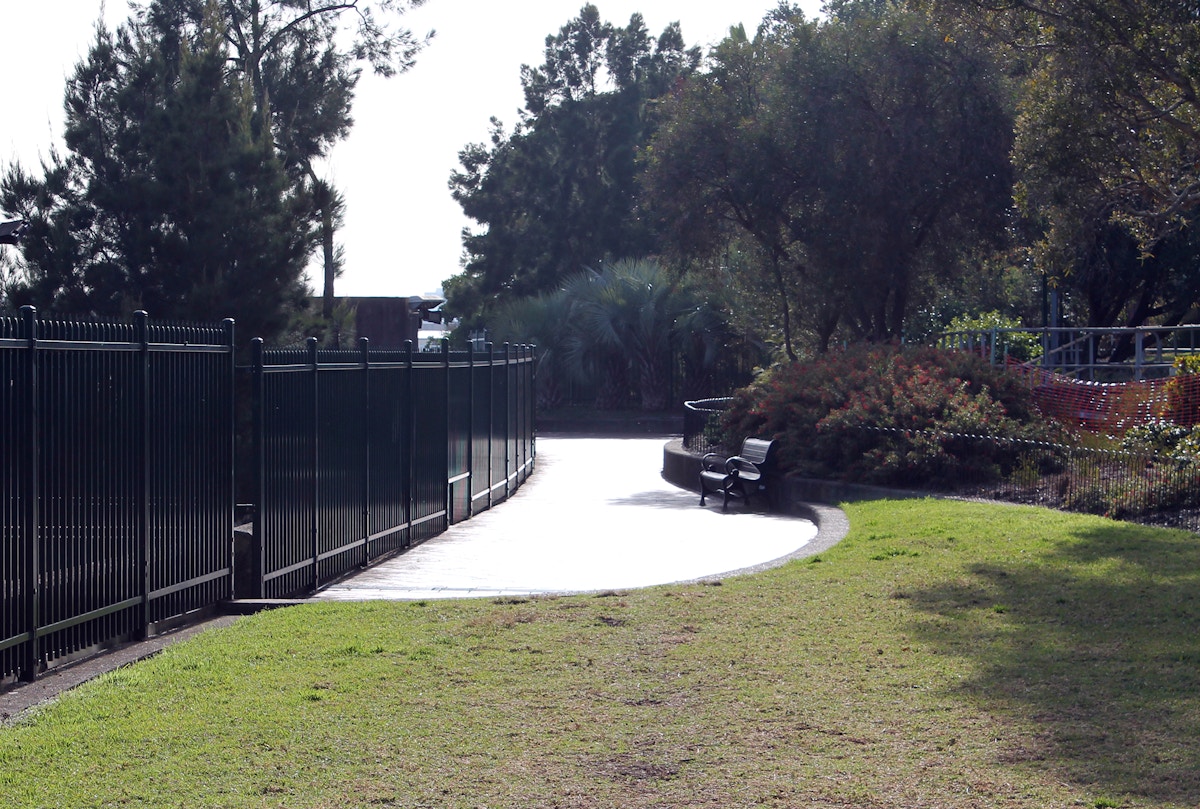 A paved path curves beside a black metal fence, with a bench and green grass on the right, surrounded by trees and bushes under daylight.