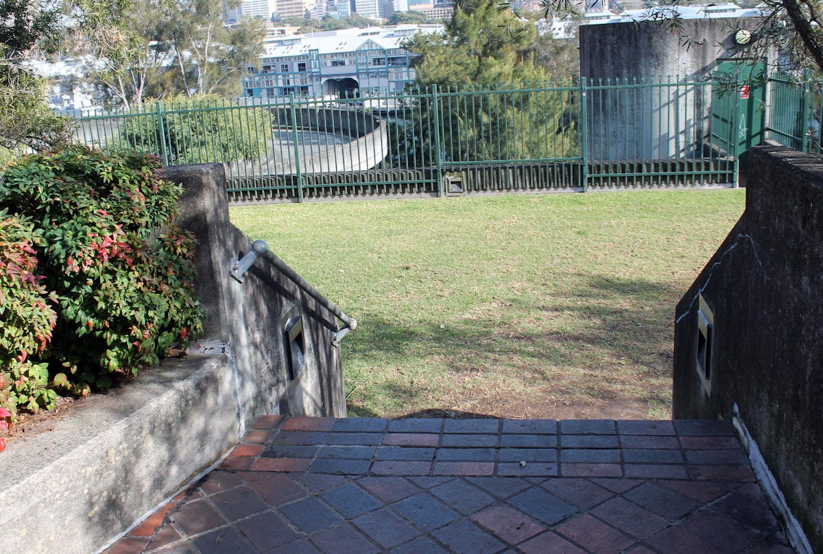 Brick steps descend to a grassy area bordered by shrubs and a metal fence, with buildings visible in the background.