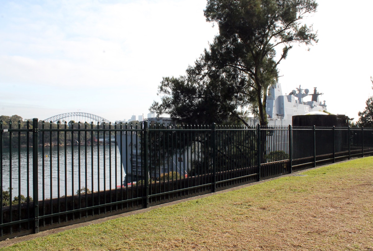 A black metal fence borders a grassy area overlooking water, with a large ship and the Sydney Harbour Bridge visible in the background.