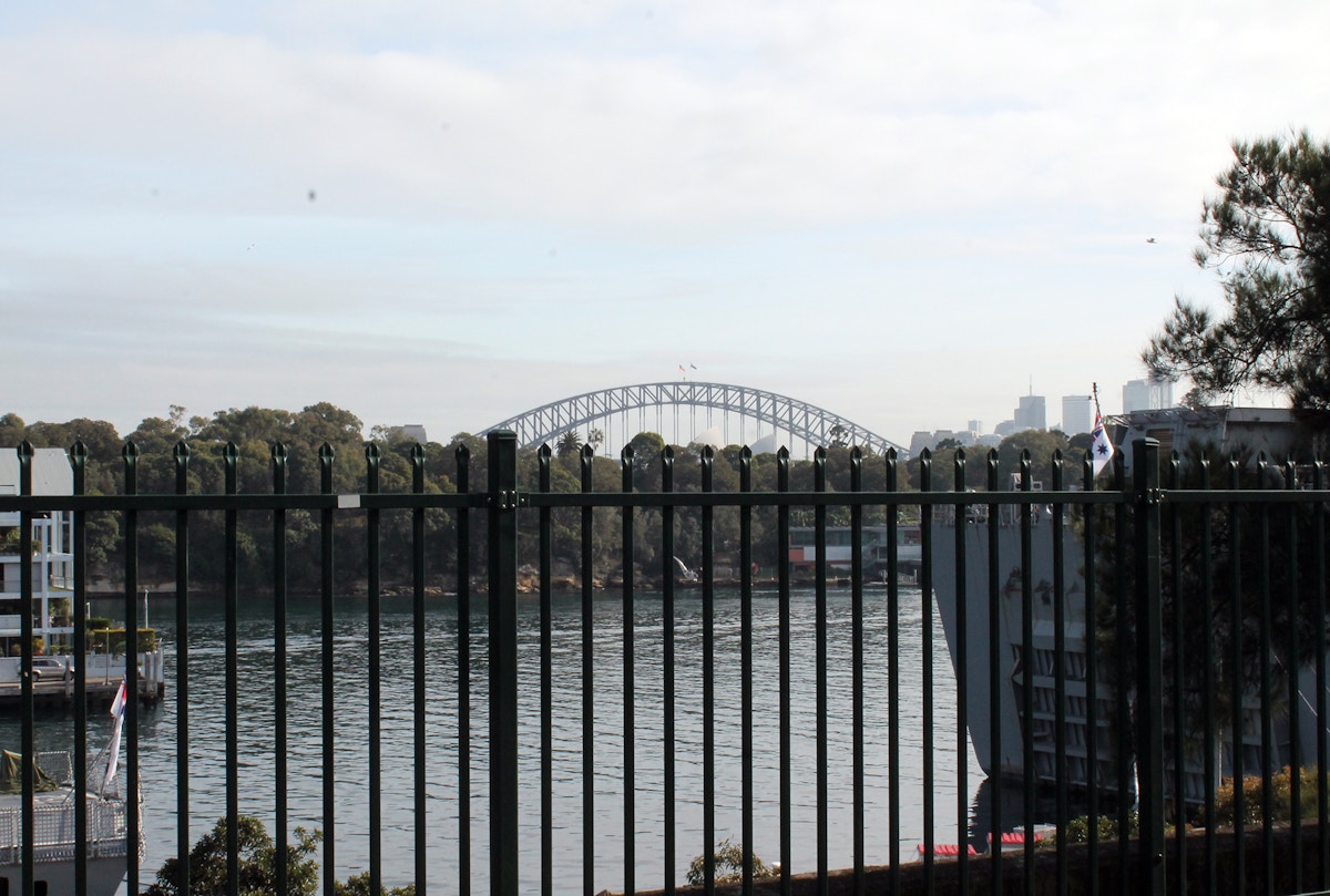 A metal fence in the foreground overlooks a body of water, with Sydney Harbour Bridge visible in the background under a cloudy sky.
