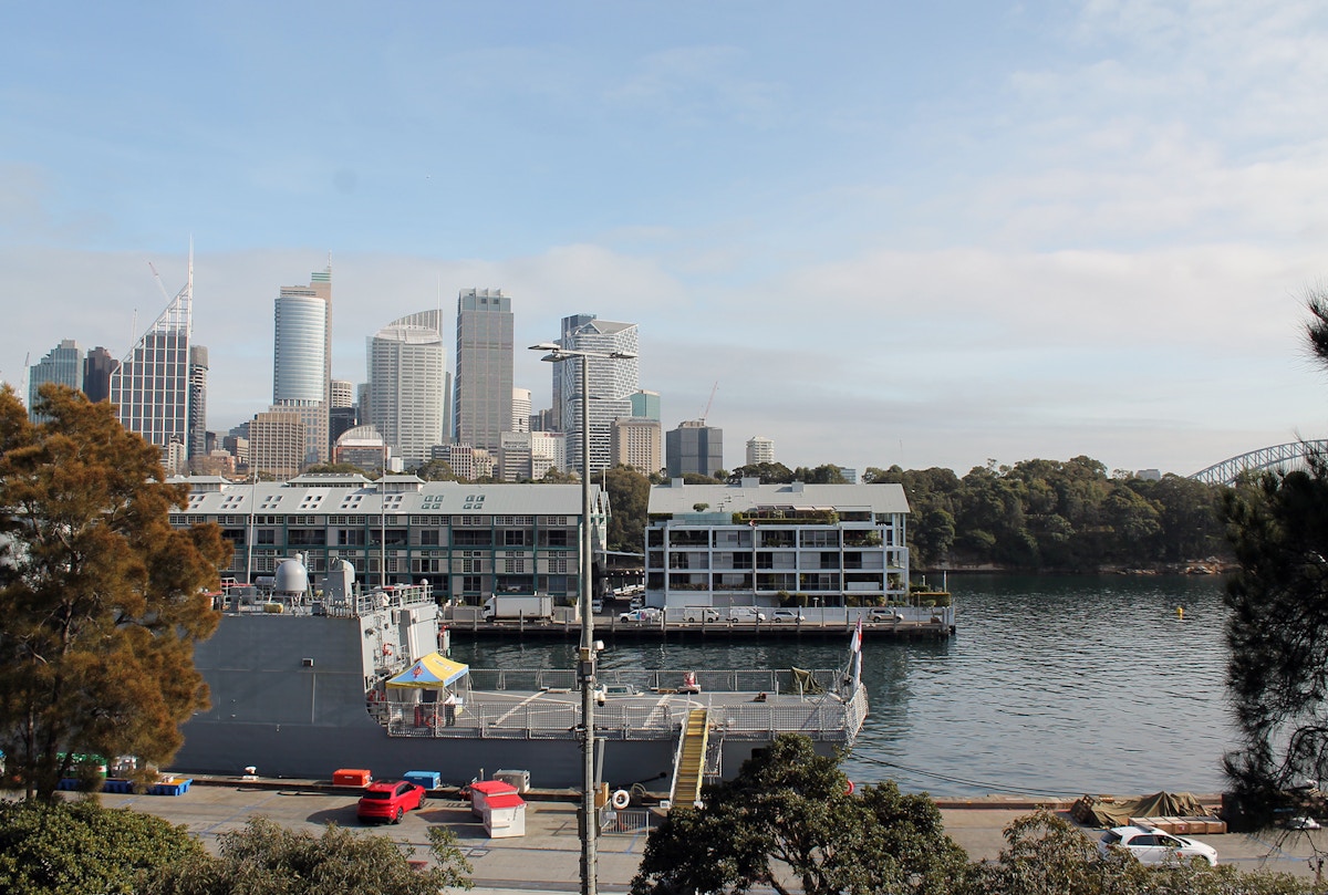 A docked navy ship is in the foreground with city skyscrapers and waterfront buildings in the background under a partly cloudy sky.