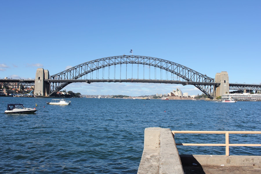 Sydney Harbour Bridge spans across the water with the Sydney Opera House visible in the background; boats are floating in the harbor under a clear blue sky.