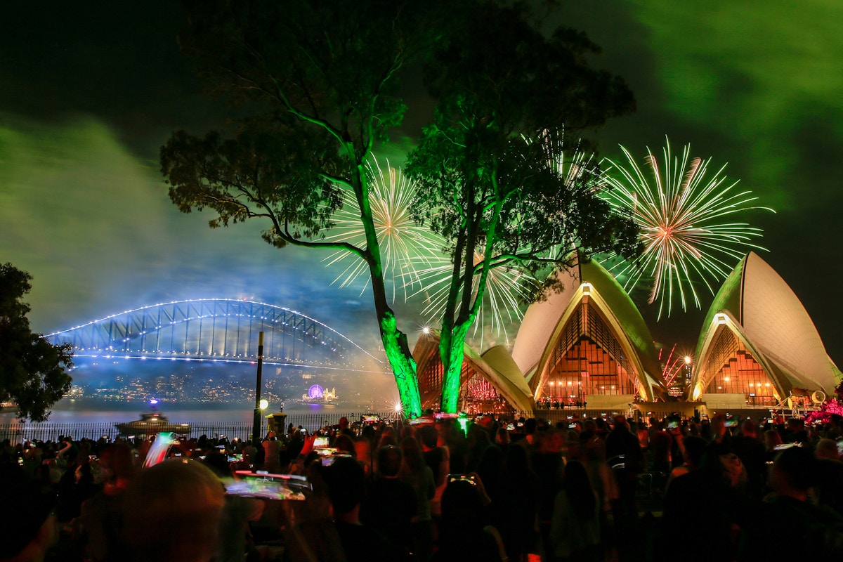 Fireworks light up the sky above Sydney Opera House and Sydney Harbour Bridge at night, with crowds watching and trees partially visible in the foreground.
