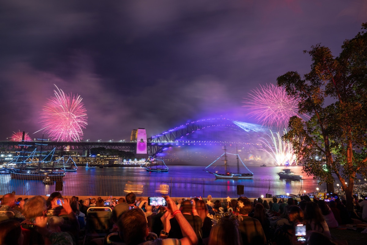 Crowd watches a waterfront fireworks display at night with Sydney Harbour Bridge illuminated in the background and several boats on the water.