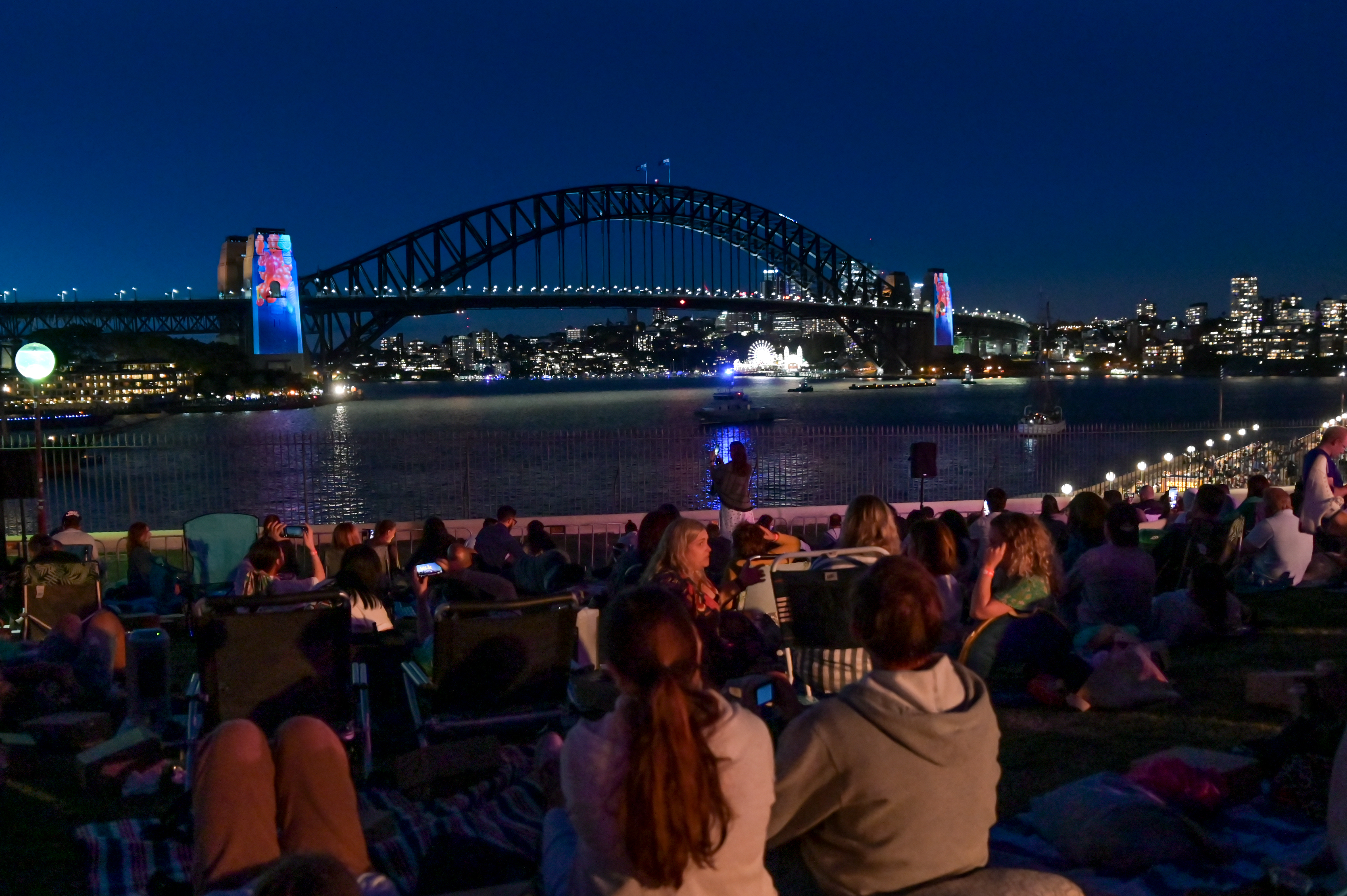People sit on a grassy area at night, facing Sydney Harbour Bridge, with city lights and illuminated bridge pylons in the background.